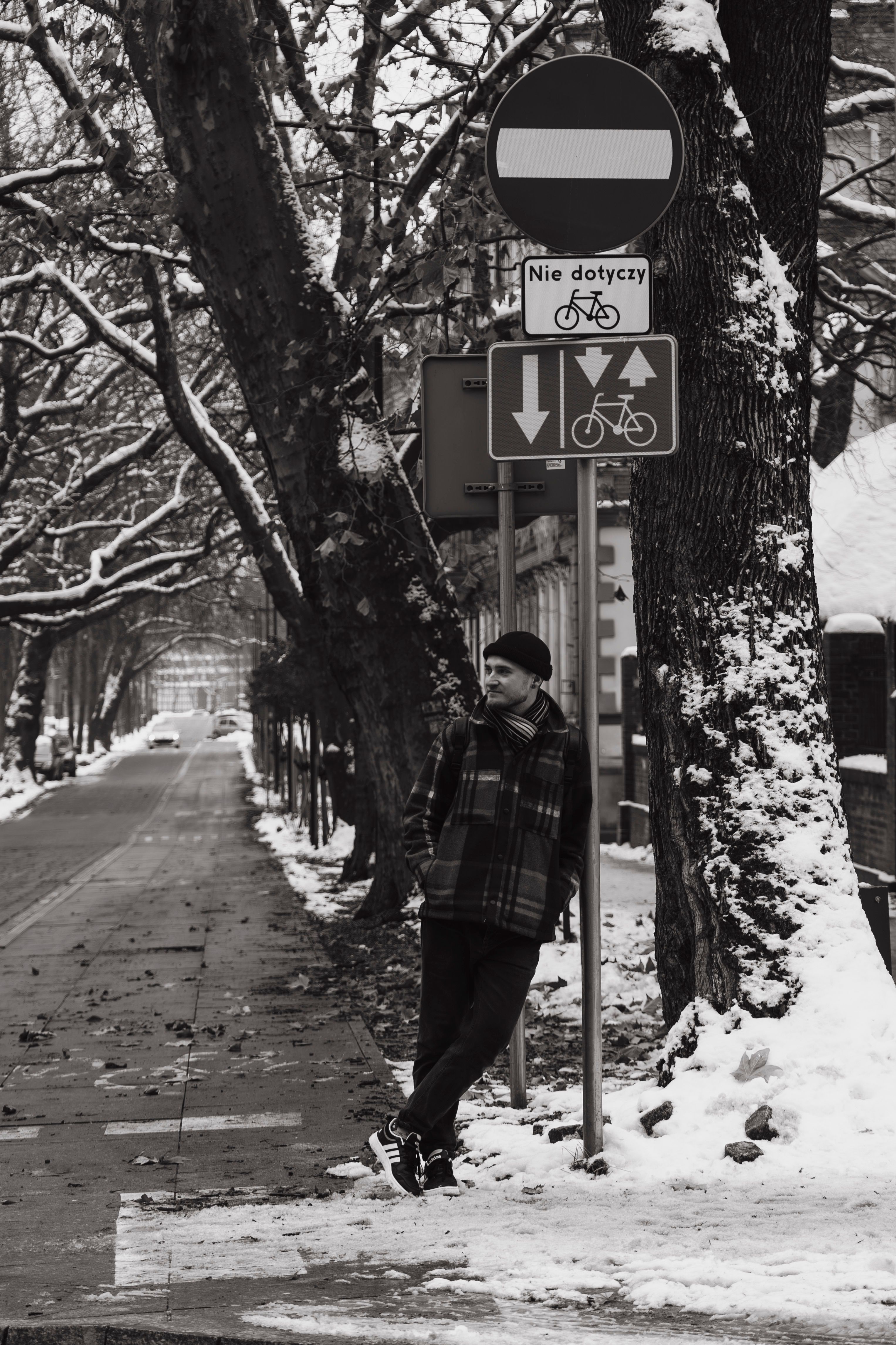 Black and white photo of a man leaning on a street sign in a snowy urban setting.
