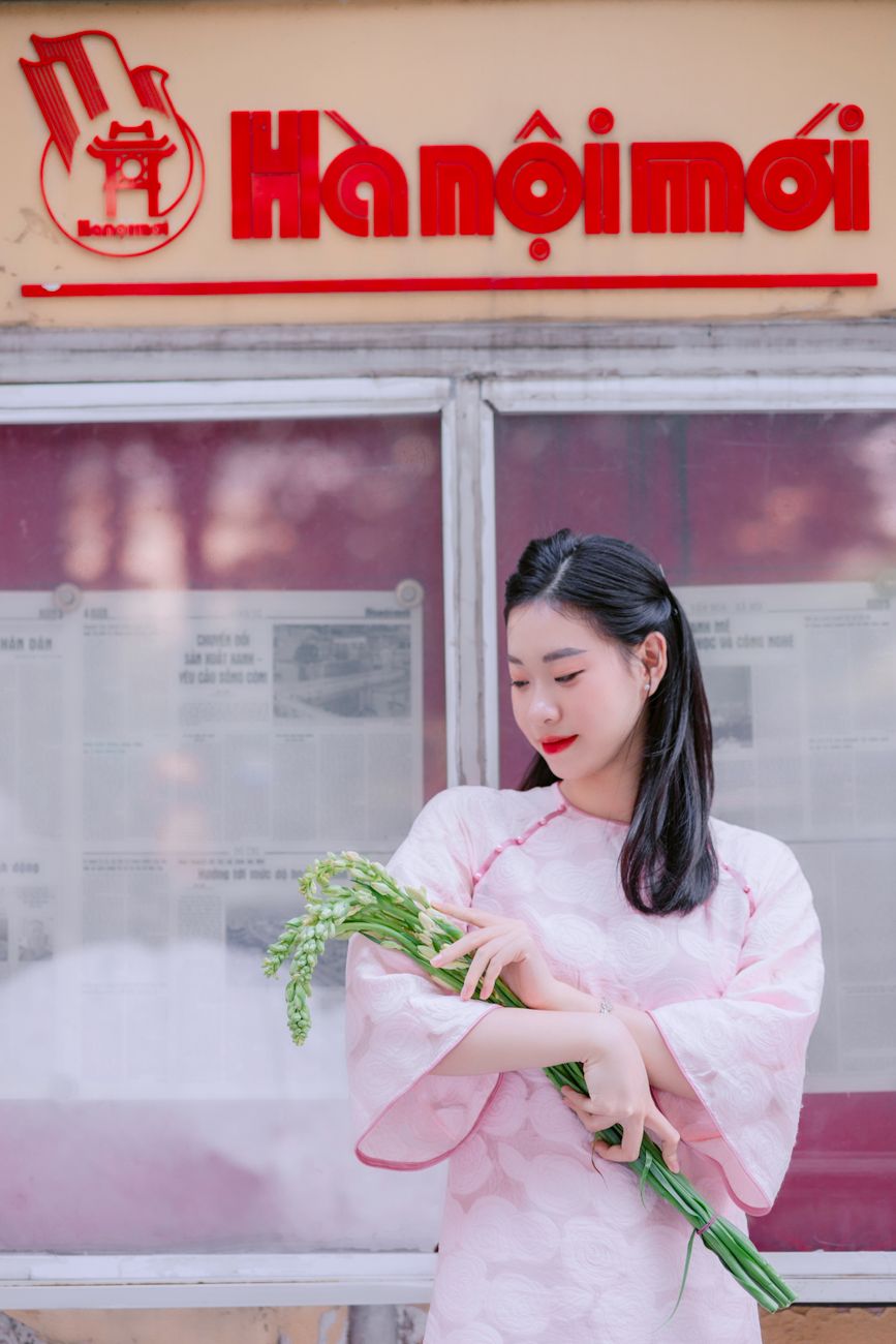 Elegant Vietnamese woman in traditional attire holding flowers near Hanoi sign, embodying cultural grace.