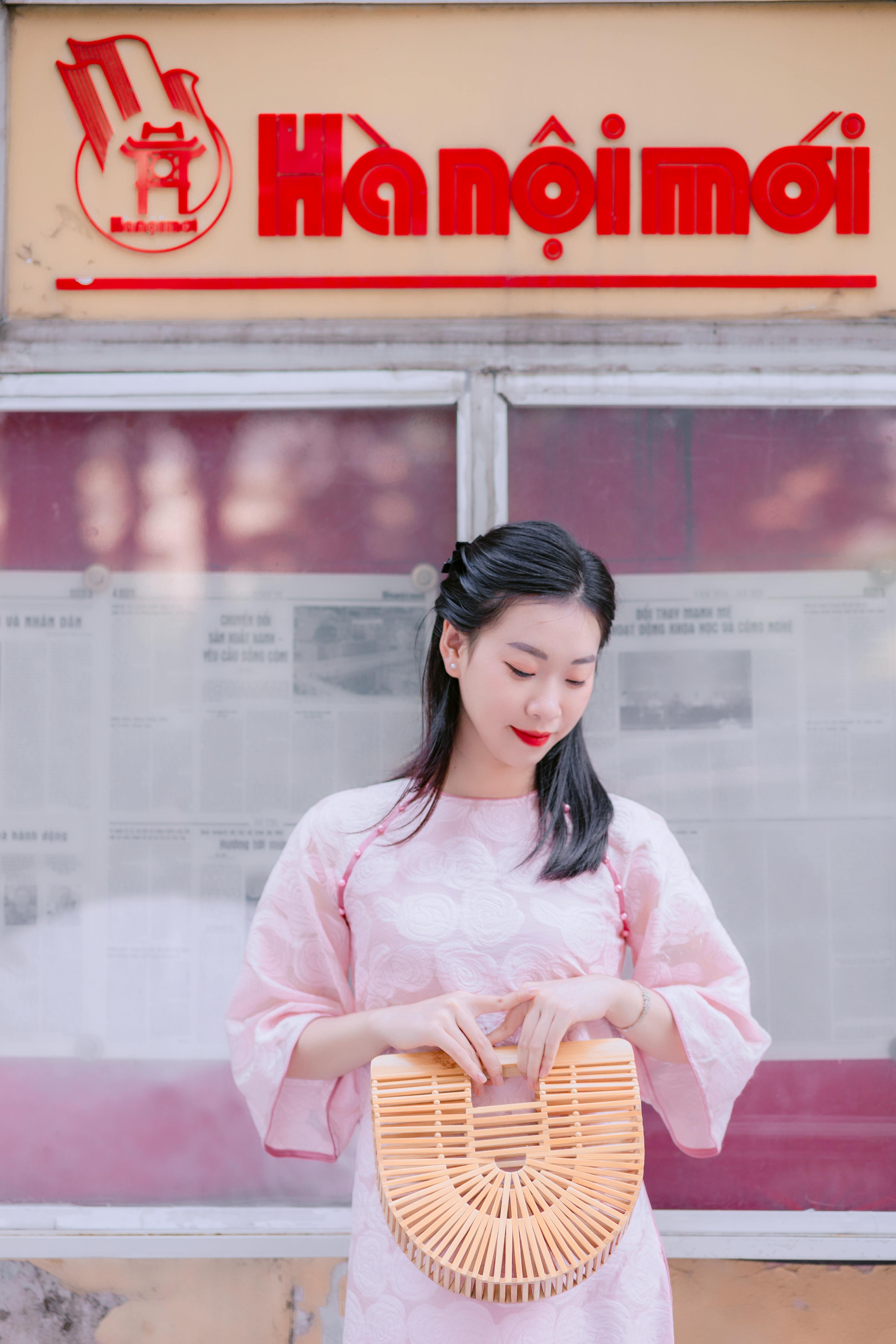 Young Vietnamese woman in traditional pink ao dai holding a bamboo basket in front of Ha Noi Moi new
