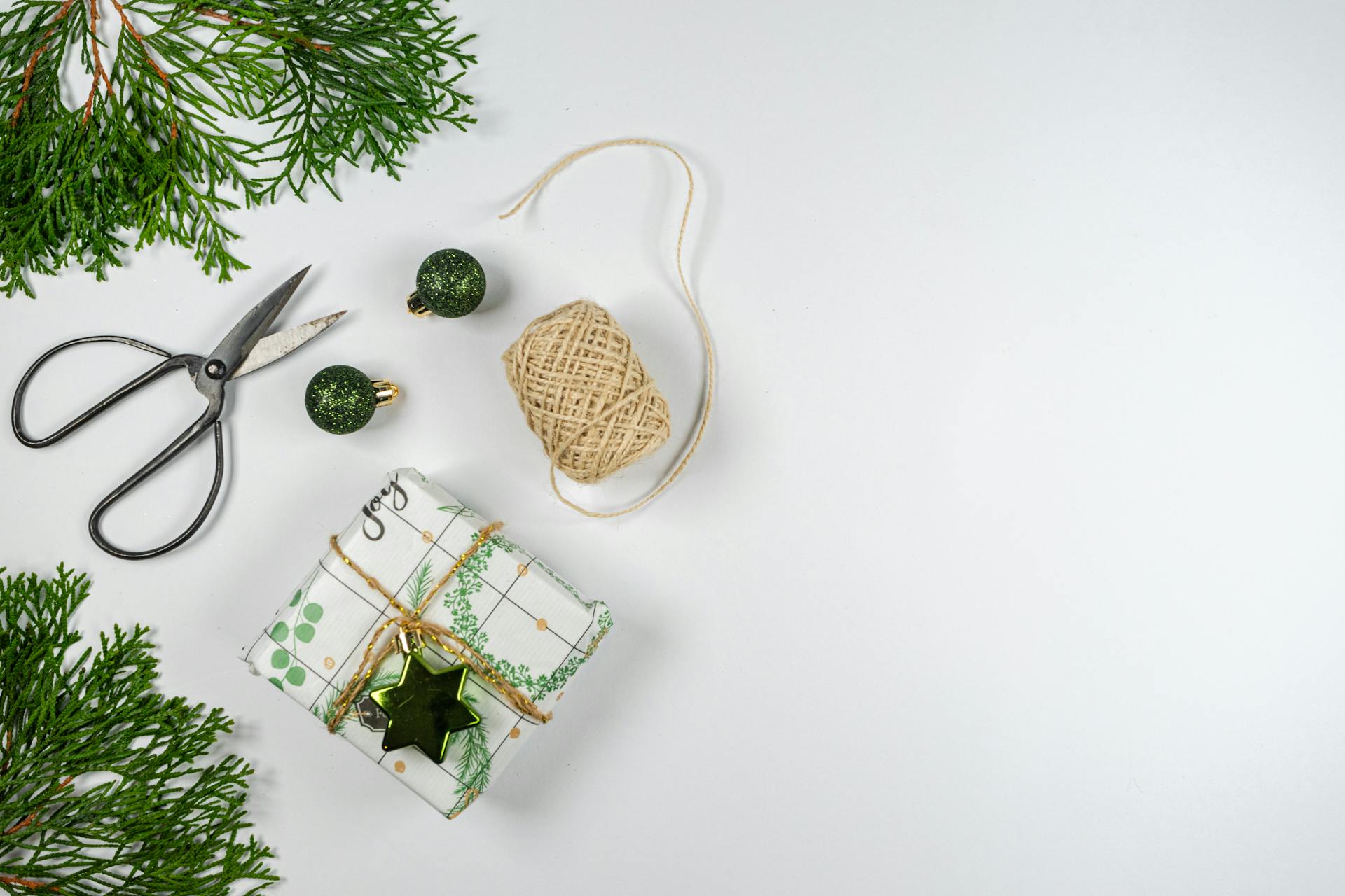 Christmas gift wrapping scene with scissors, twine, and green decorations on a white background.