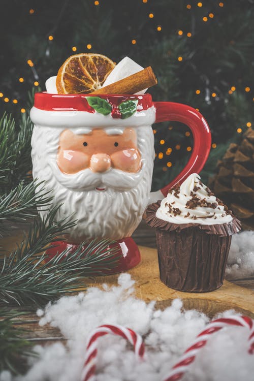 Close-up of a Drink in a Santa Claus Mug Standing next to a Cupcake on the Background of a Christmas Tree