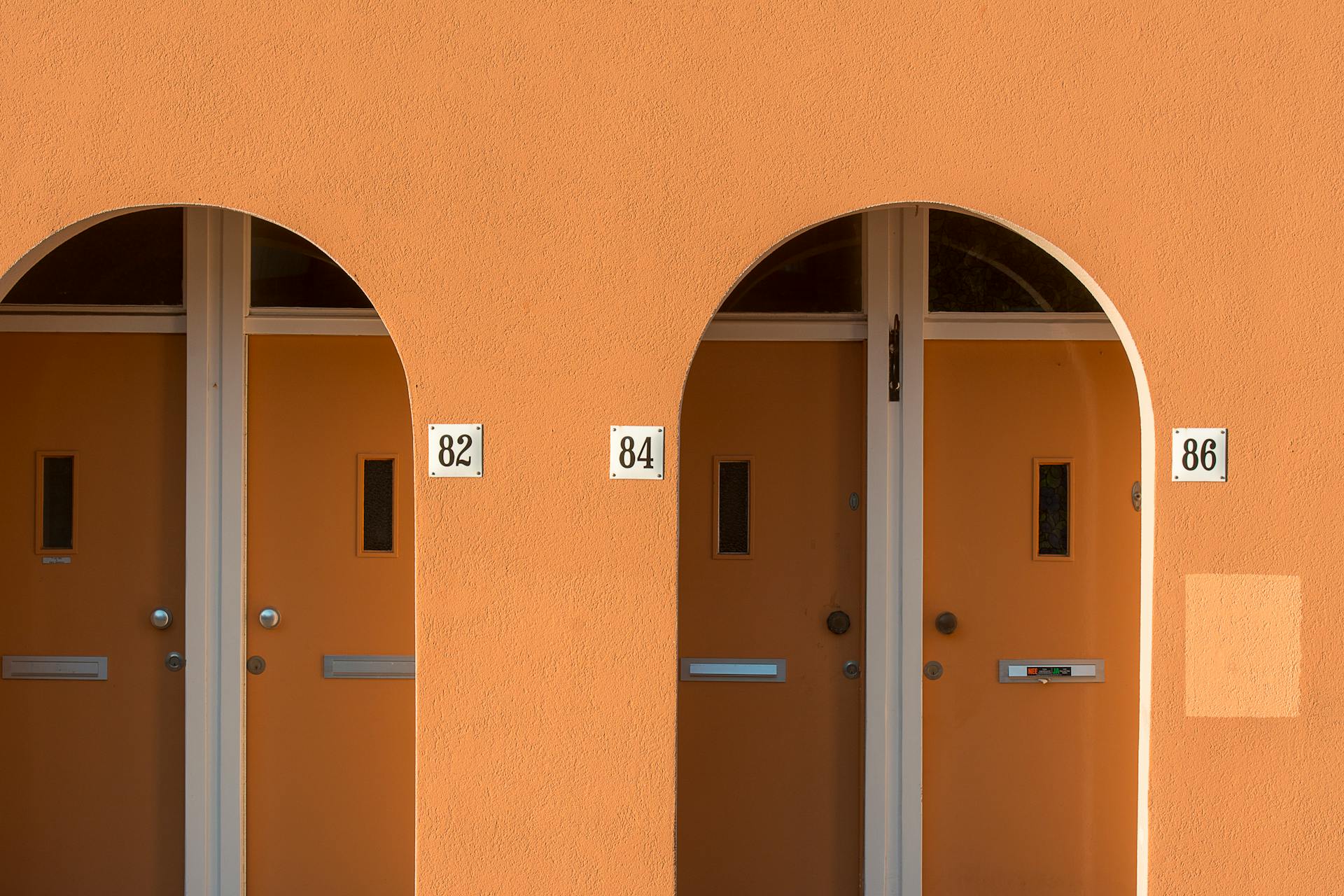 Stylish orange doors with arched frames and house numbers 82, 84, 86 on a sunny wall.