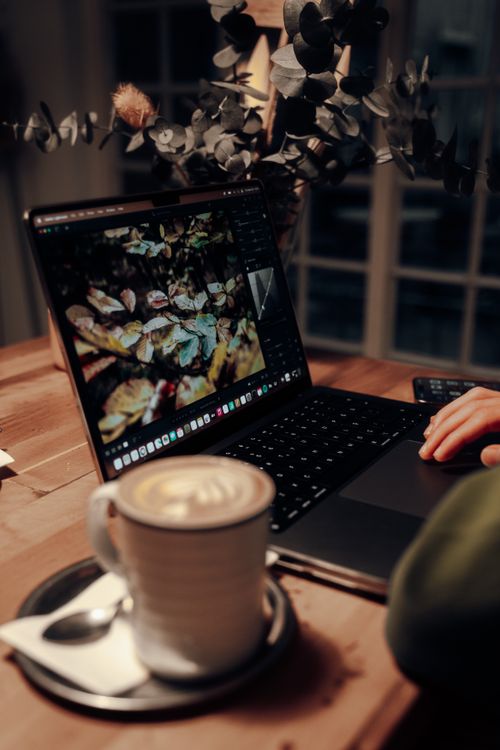 Free A cozy indoor workspace featuring a laptop and a steaming cup of cappuccino on a wooden desk. Stock Photo