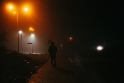 Free A lone silhouette walks on a foggy street at night, illuminated by streetlamps, creating a moody atmosphere. Stock Photo