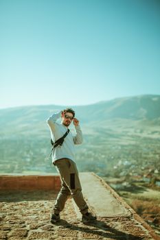 A fashionable man adjusting glasses on a rooftop with a scenic cityscape view.