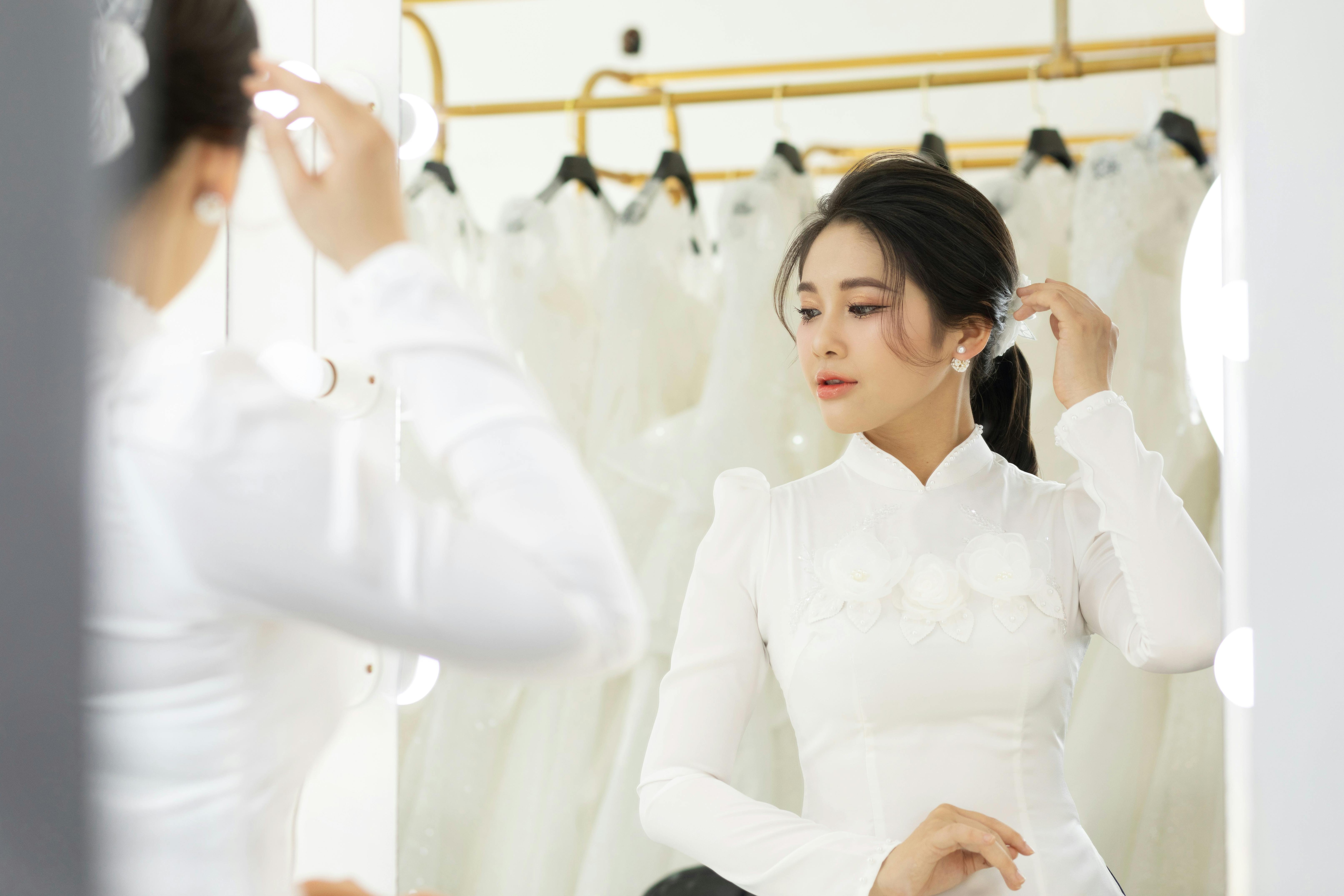 A beautiful bride in a white dress adjusts her hair in front of a mirror in a bridal shop.