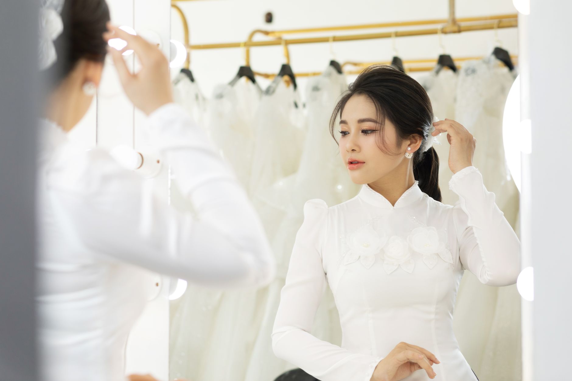 A bride in a white gown adjusts her hair in front of a mirror in a bridal shop.