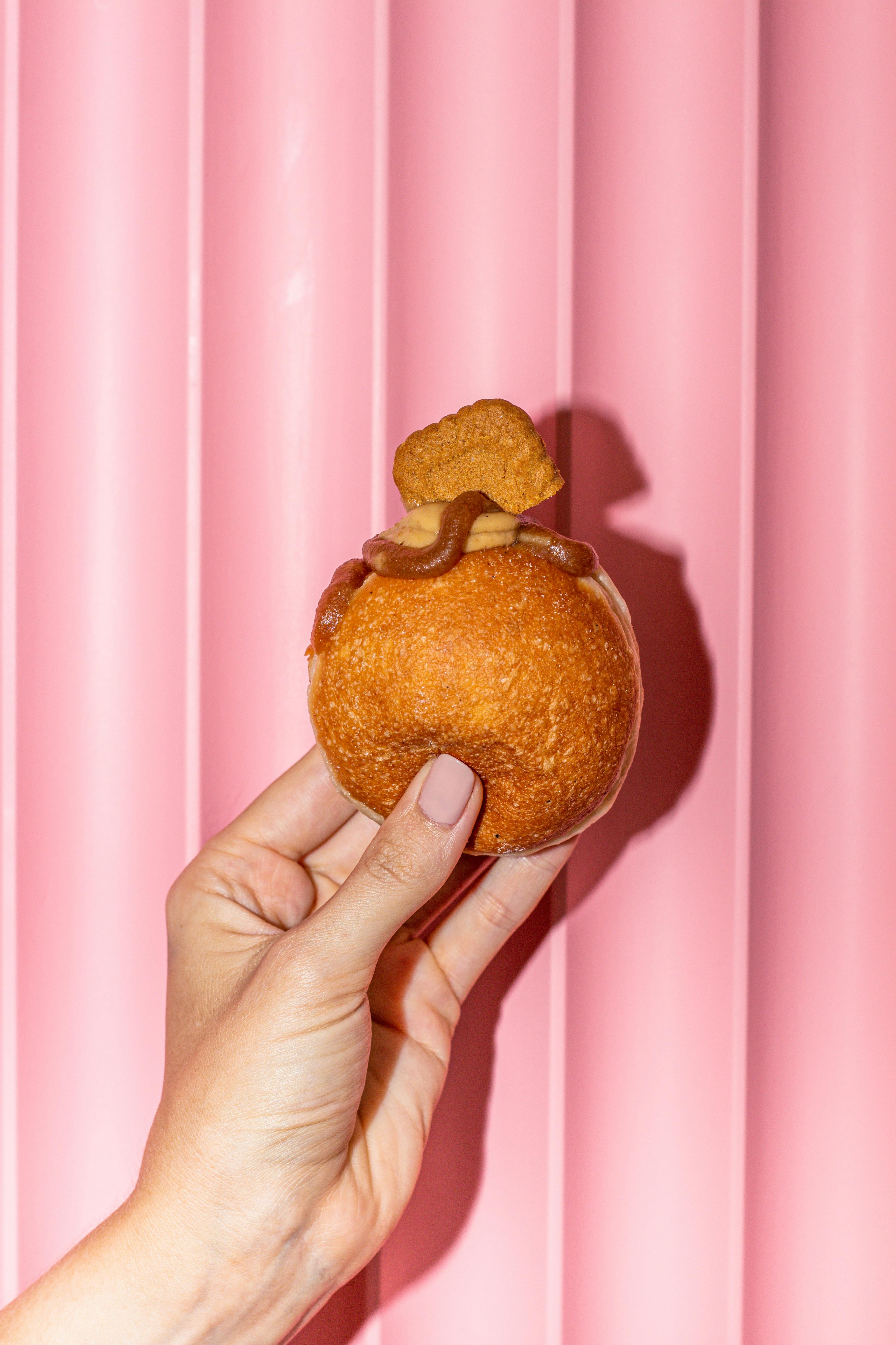 A hand holds a donut topped with caramel and cookie against a pink backdrop, perfect for snack concepts.