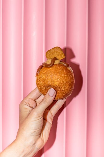 A hand holds a donut topped with caramel and cookie against a pink backdrop, perfect for snack concepts.