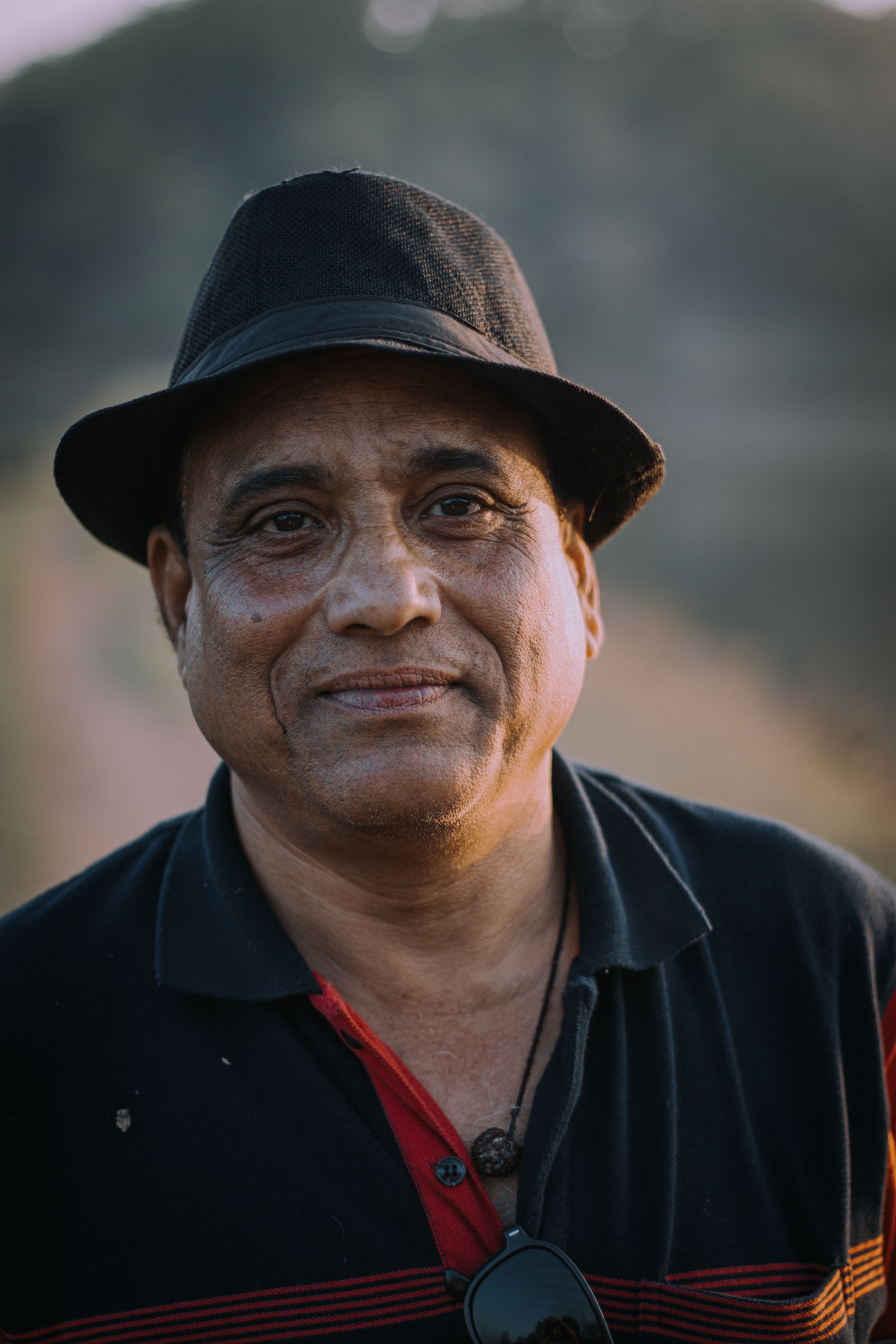 A close-up portrait of a mature man in a black fedora and polo shirt shot outdoors.