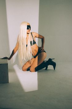 Blonde woman in black lingerie and mask posing in a studio setting.