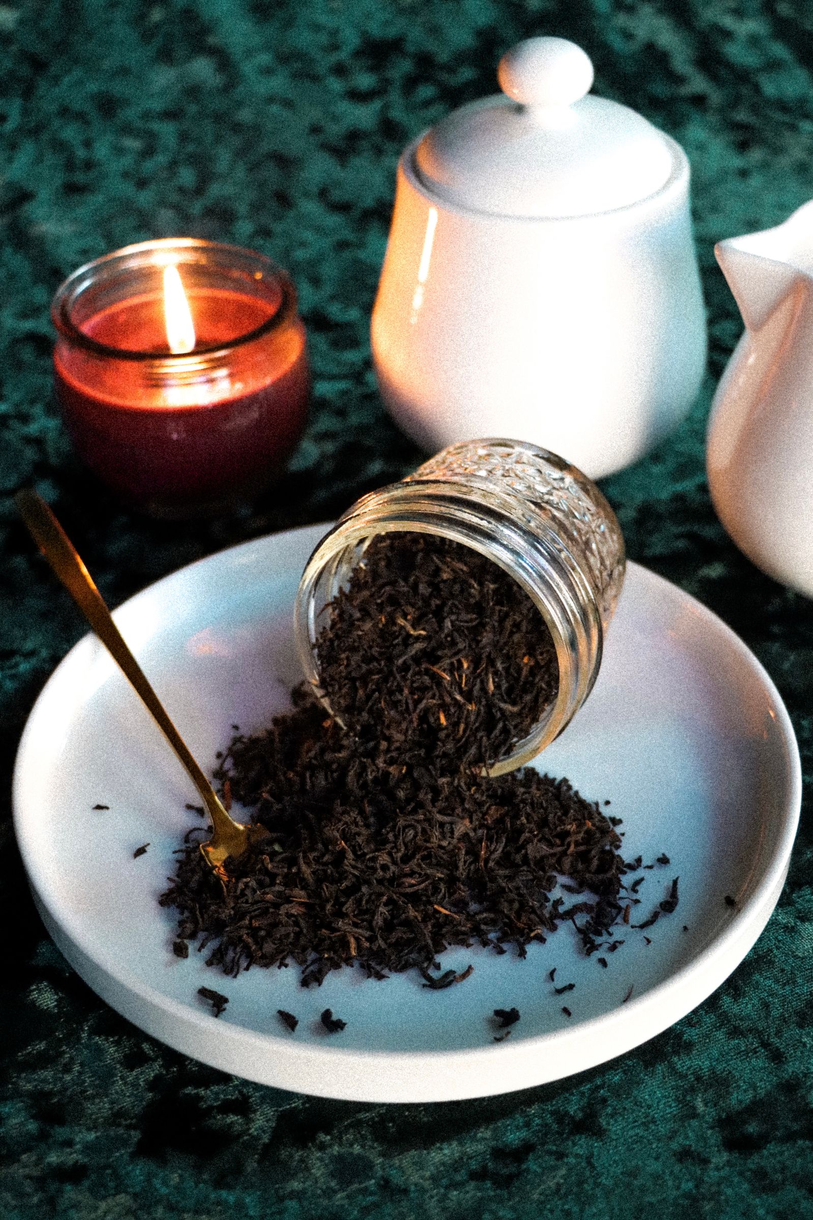 Tea leaves spilling from a jar beside a candle and dark teapot