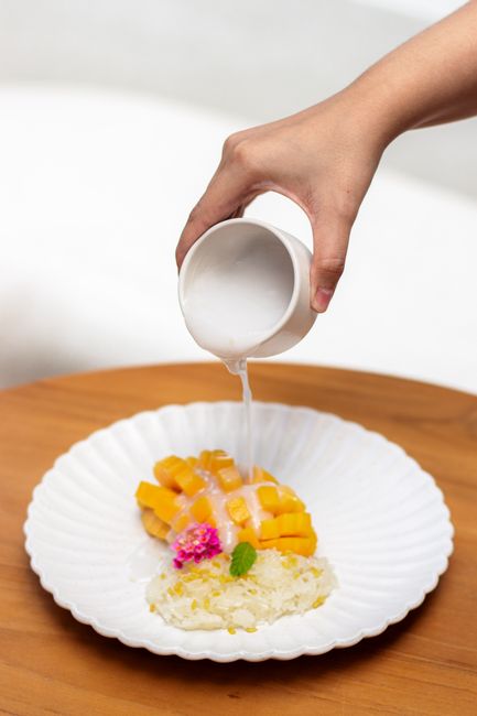 Hand pouring coconut milk over traditional Thai mango sticky rice dessert.
