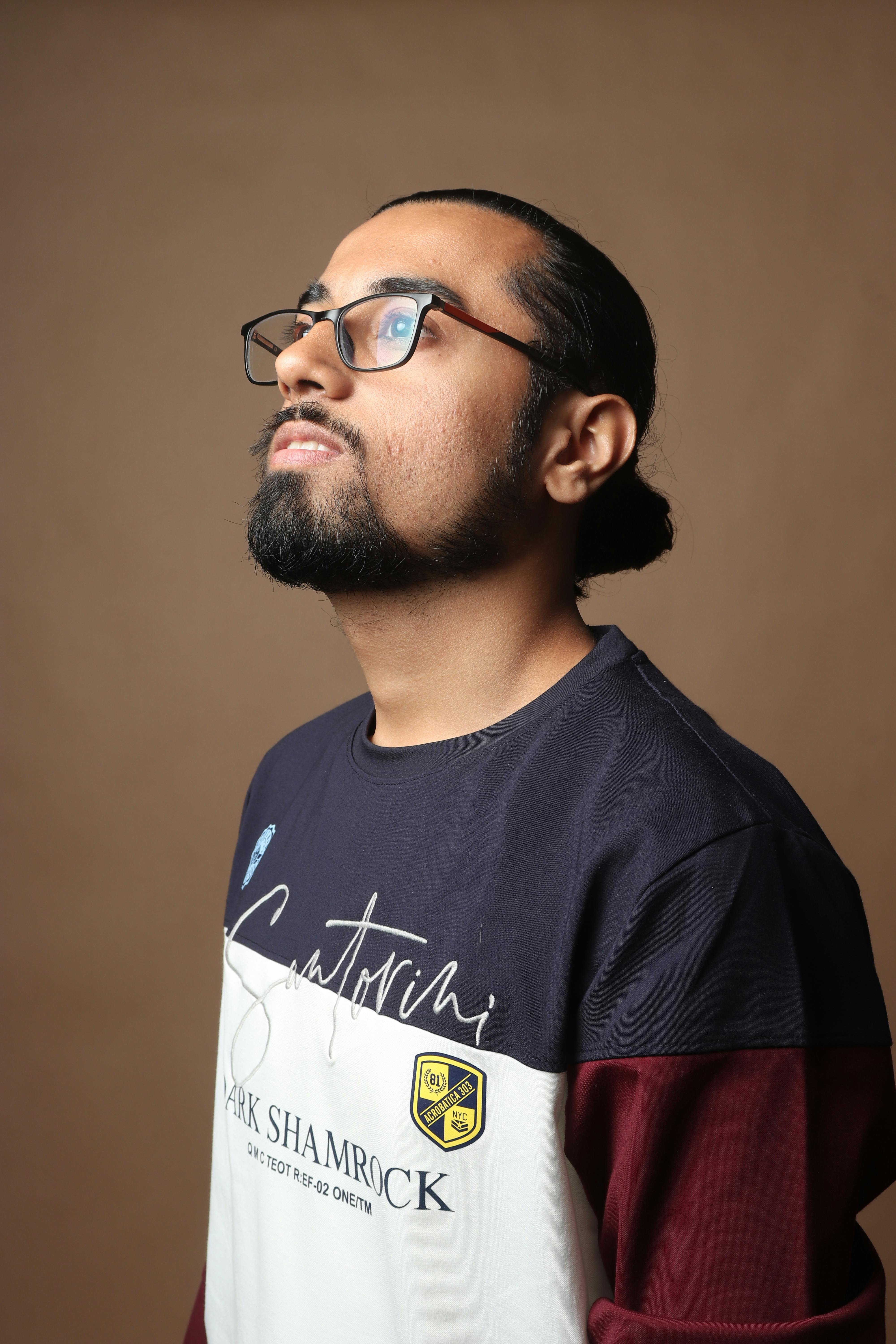 A contemplative bearded man gazing upward, wearing eyeglasses and casual wear against a brown backdrop.
