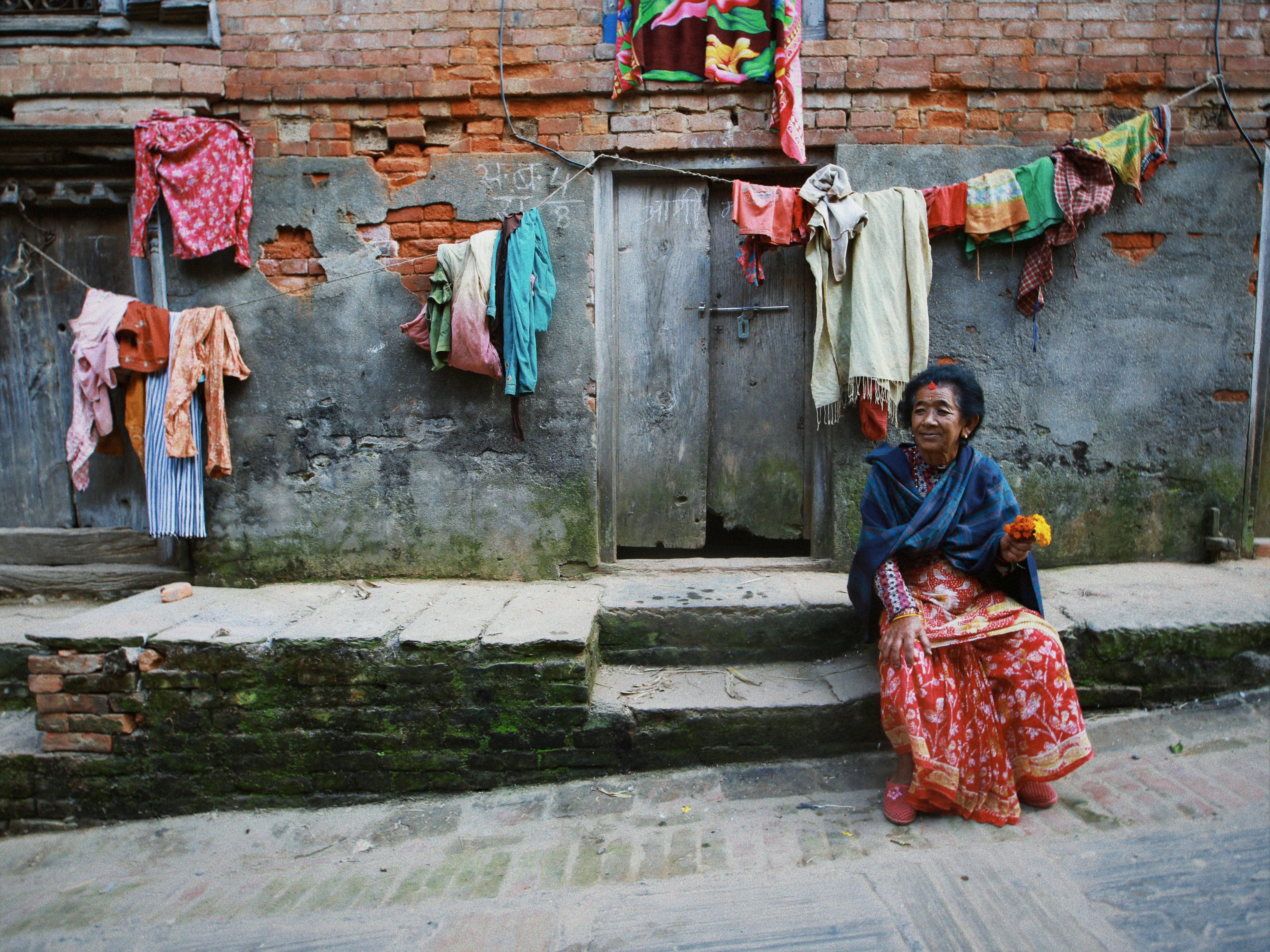 An elderly woman sits on steps next to colorful hanging laundry on a rustic street.