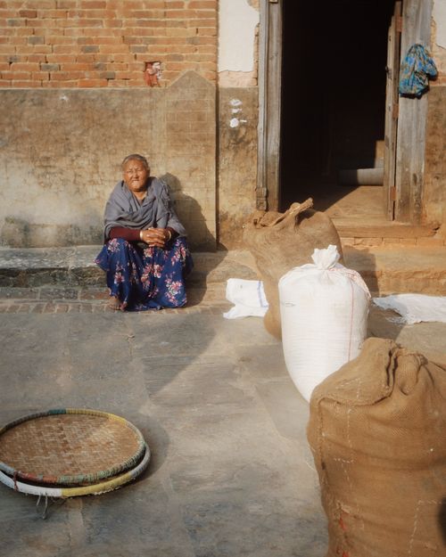 Free An elderly woman sits outside a rustic building with sacks and baskets around her, under warm daylight. Stock Photo