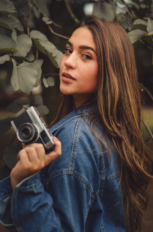 Woman Wearing Blue Denim Jacket While Holding Camera Near Tree