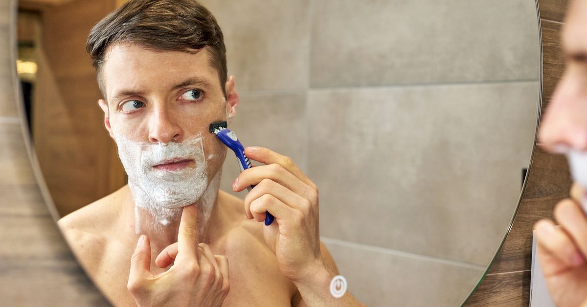 A man uses a razor for shaving in front of a bathroom mirror, focusing carefully.