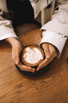 A cozy close-up of a woman holding a cappuccino with latte art on a wooden table.