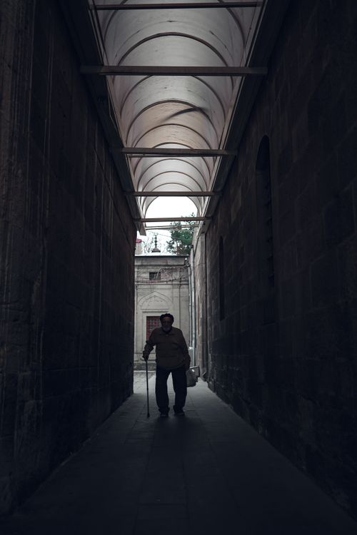 Free Silhouette of an elderly man with a cane walking through a dimly lit historic alleyway. Stock Photo