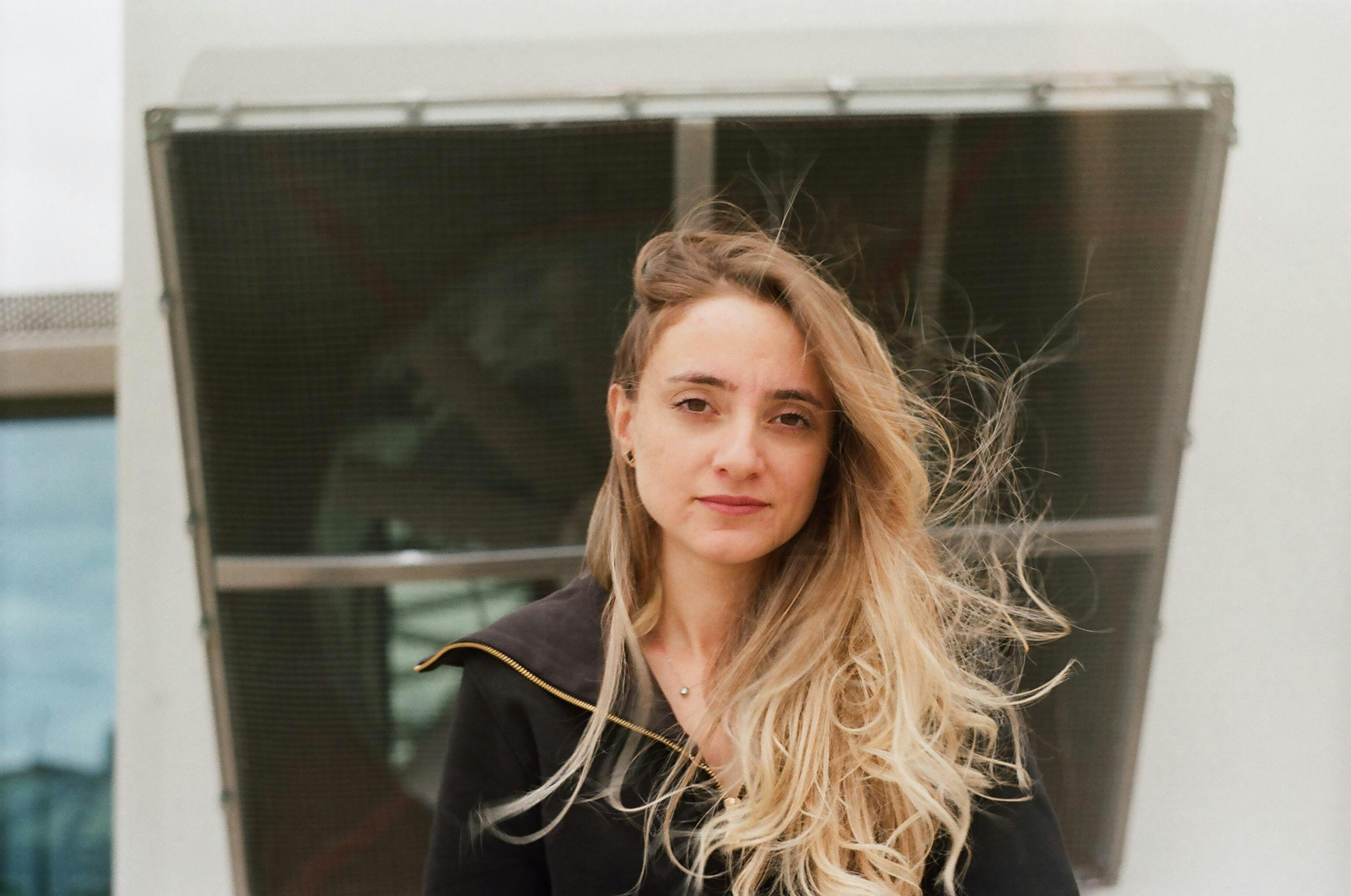 Portrait of a blonde woman standing outdoors with urban backdrop.