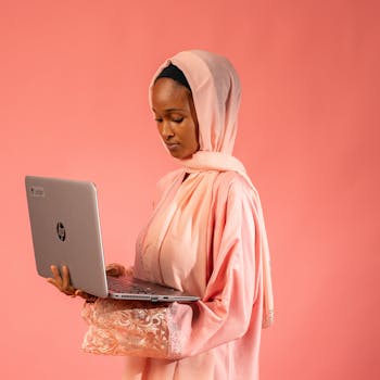 African woman in a pink headscarf working on a laptop against a pink backdrop.