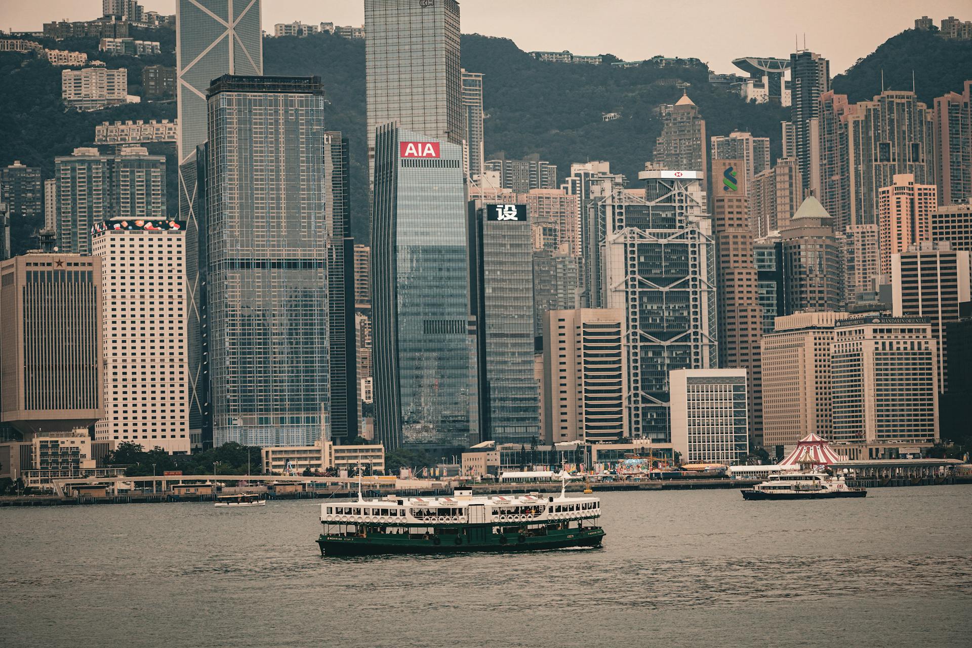 A view of Hong Kong skyscrapers with a ferry sailing on Victoria Harbor, showcasing urban architecture.