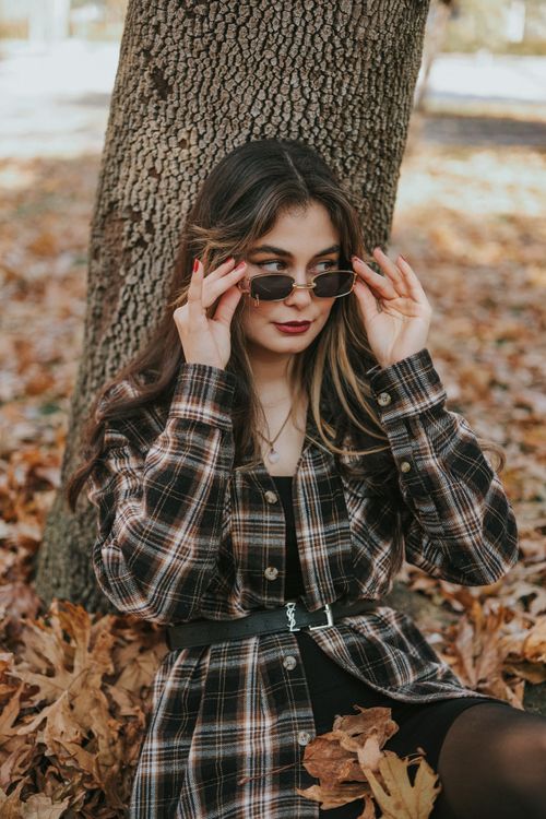 Free Young woman in checkered outfit poses with sunglasses beside a tree during fall. Stock Photo