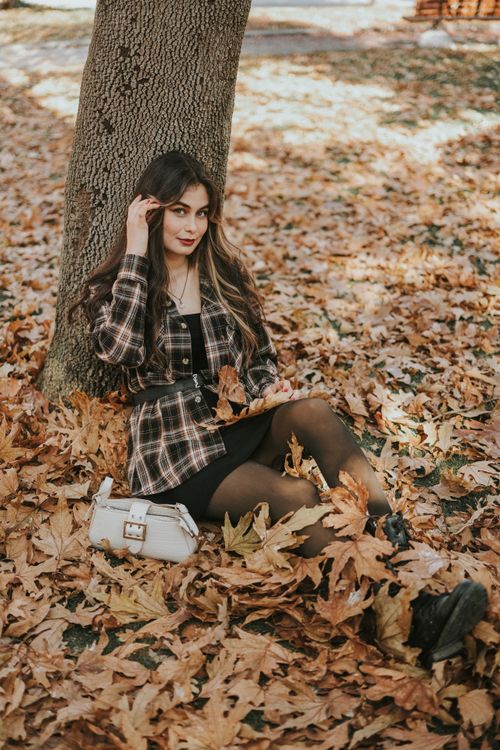 Free A stylish young woman in checkered attire sitting on autumn leaves in a park. Stock Photo