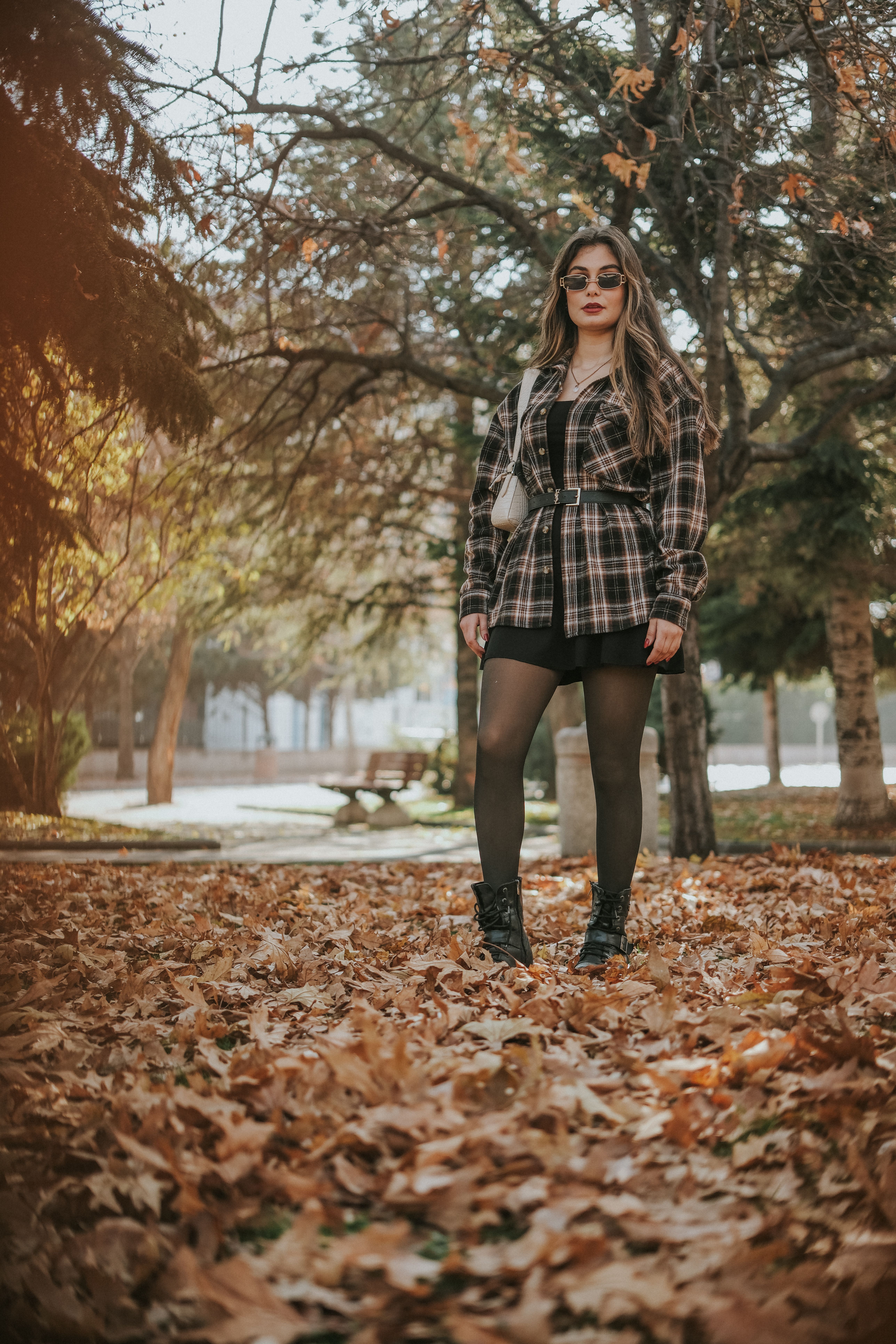 Stylish woman in sunglasses posing in autumn park, surrounded by fallen leaves on a sunny day.