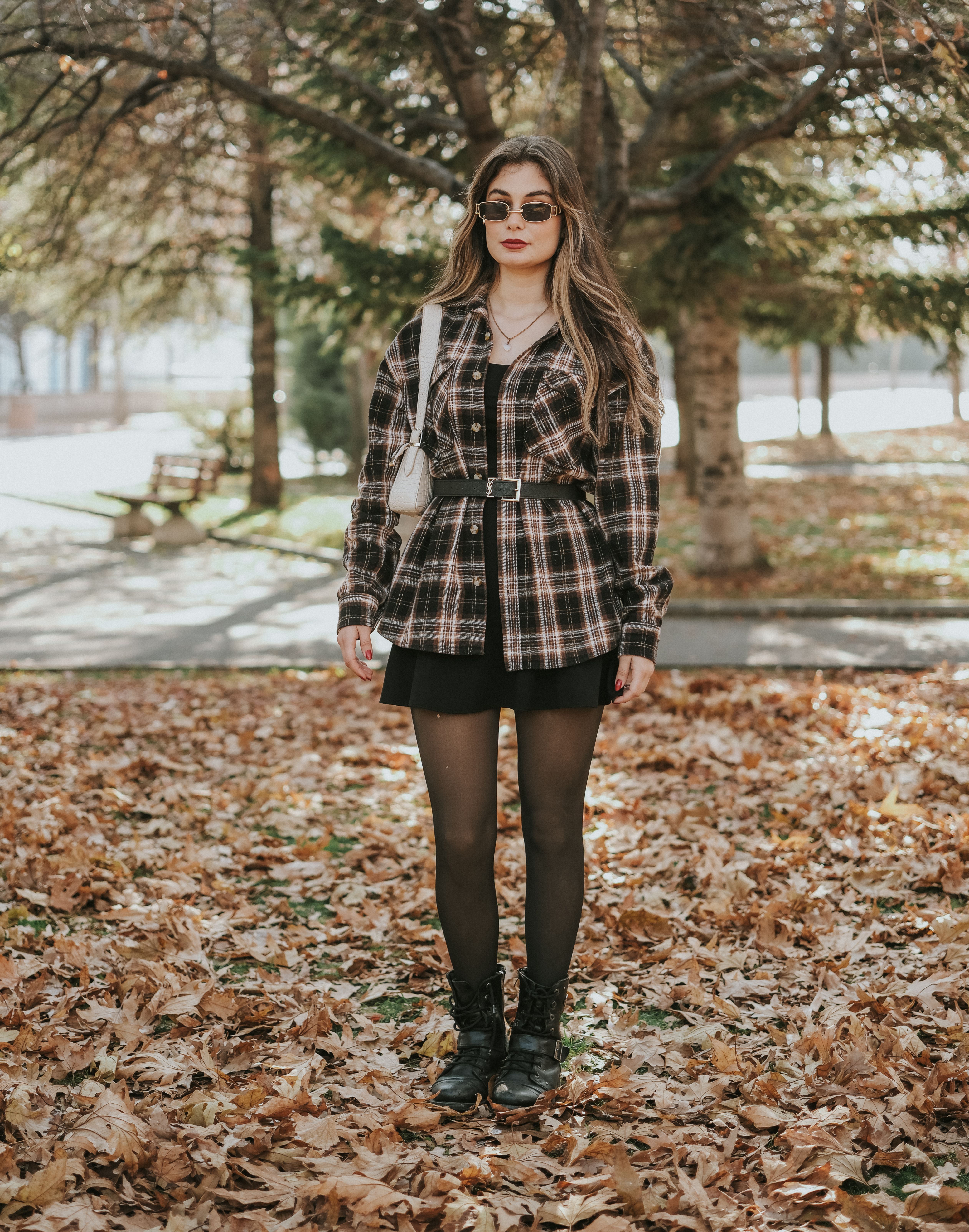 Young woman in fashionable outfit poses in a park during fall, surrounded by colorful leaves.