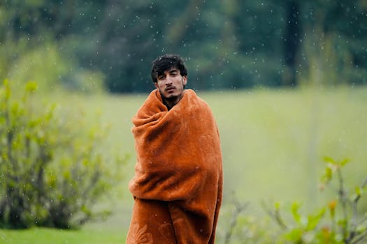 Man standing outdoors in rain, wrapped in a warm orange blanket in a lush green meadow.