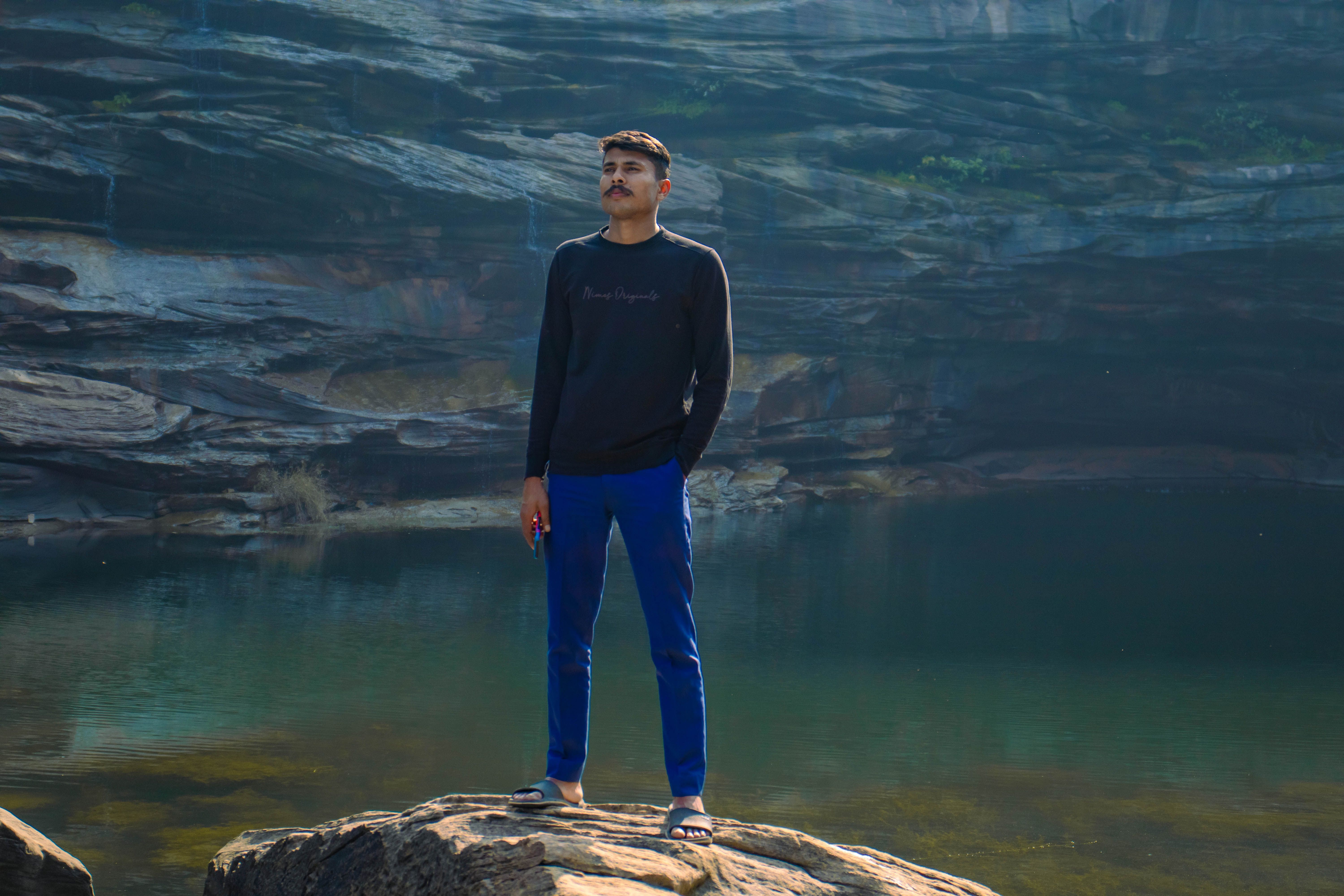 Young man standing on rocky shore by serene water on a summer day.