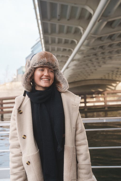 Young Woman in a Coat and Hat Standing by the River in City 