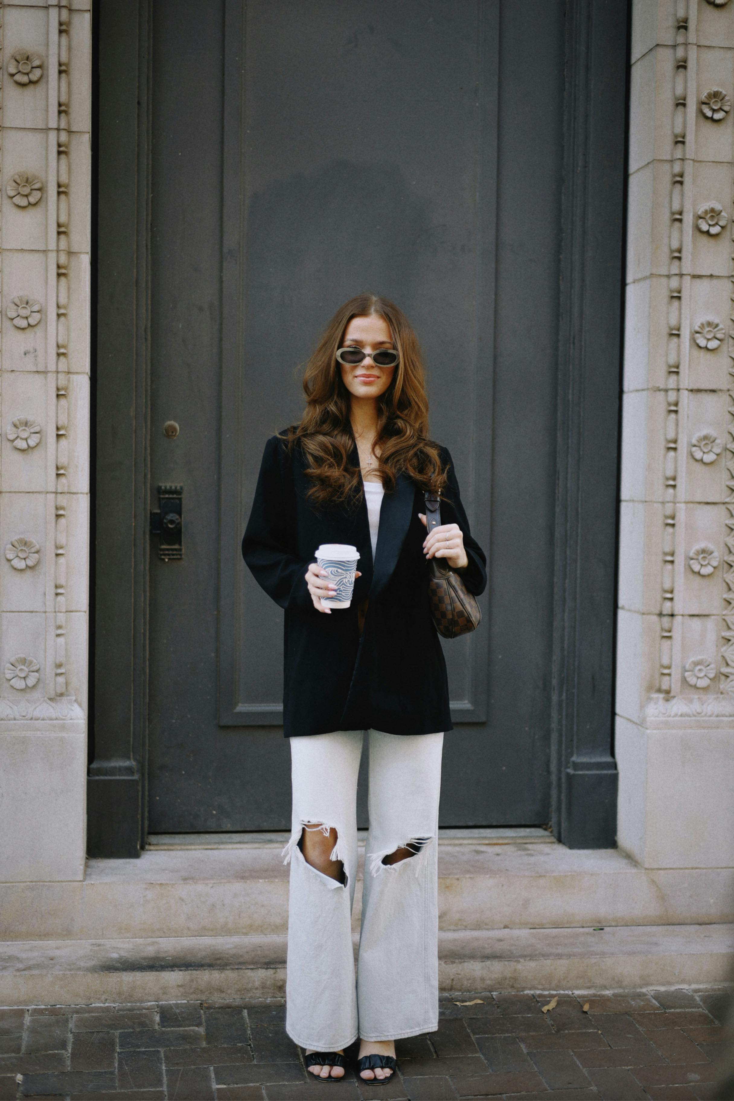 Fashionable woman with coffee in hand, wearing sunglasses, standing at an ornate door.
