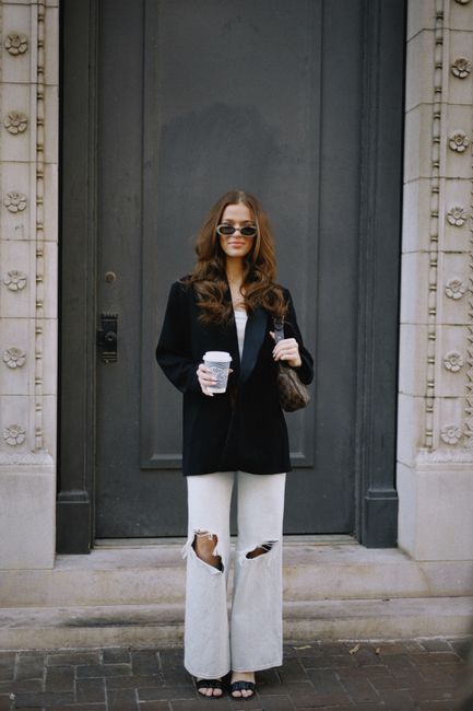 Fashionable woman with coffee in hand, wearing sunglasses, standing at an ornate door.