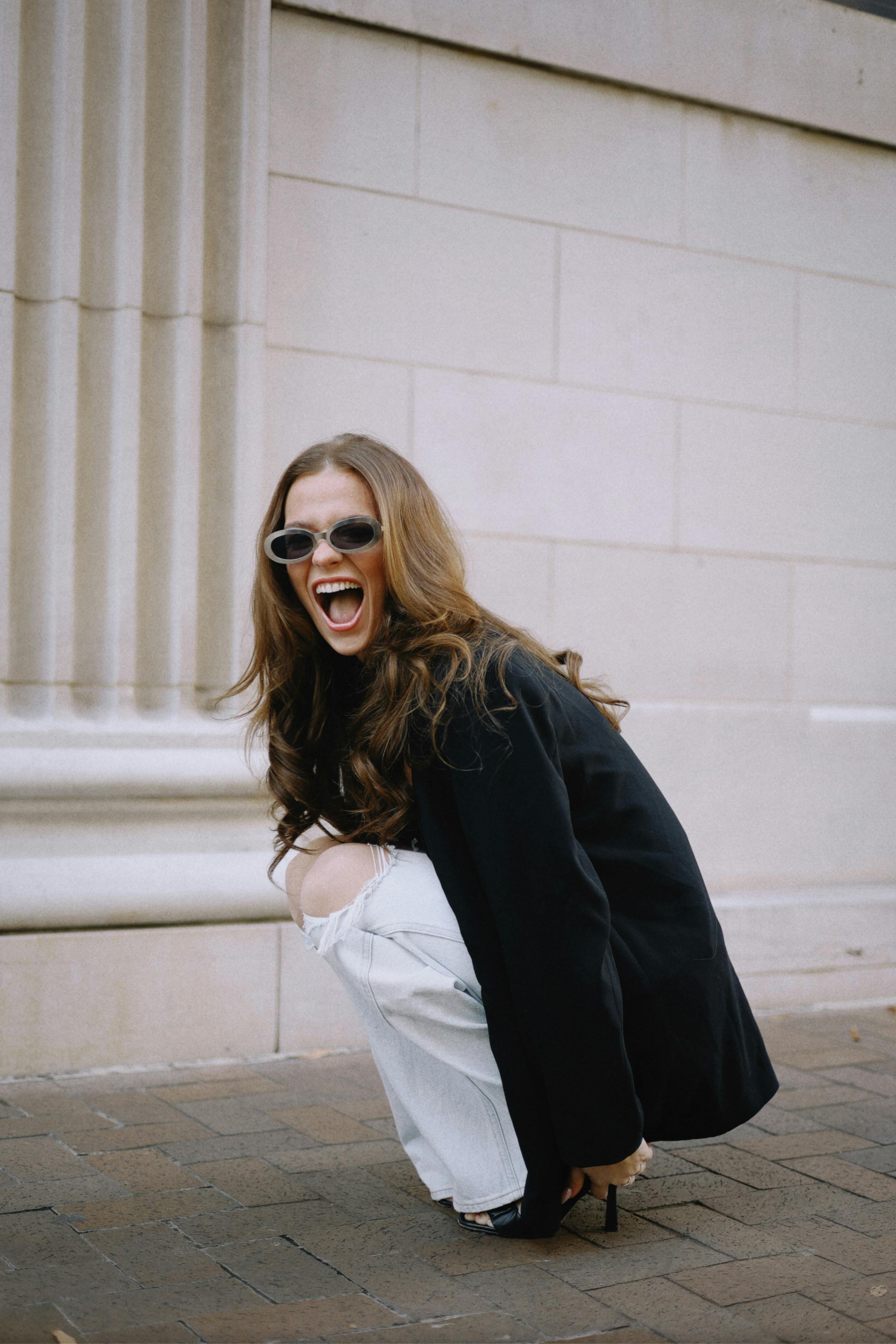 Stylish young woman in sunglasses crouching on the sidewalk, showcasing modern urban fashion.