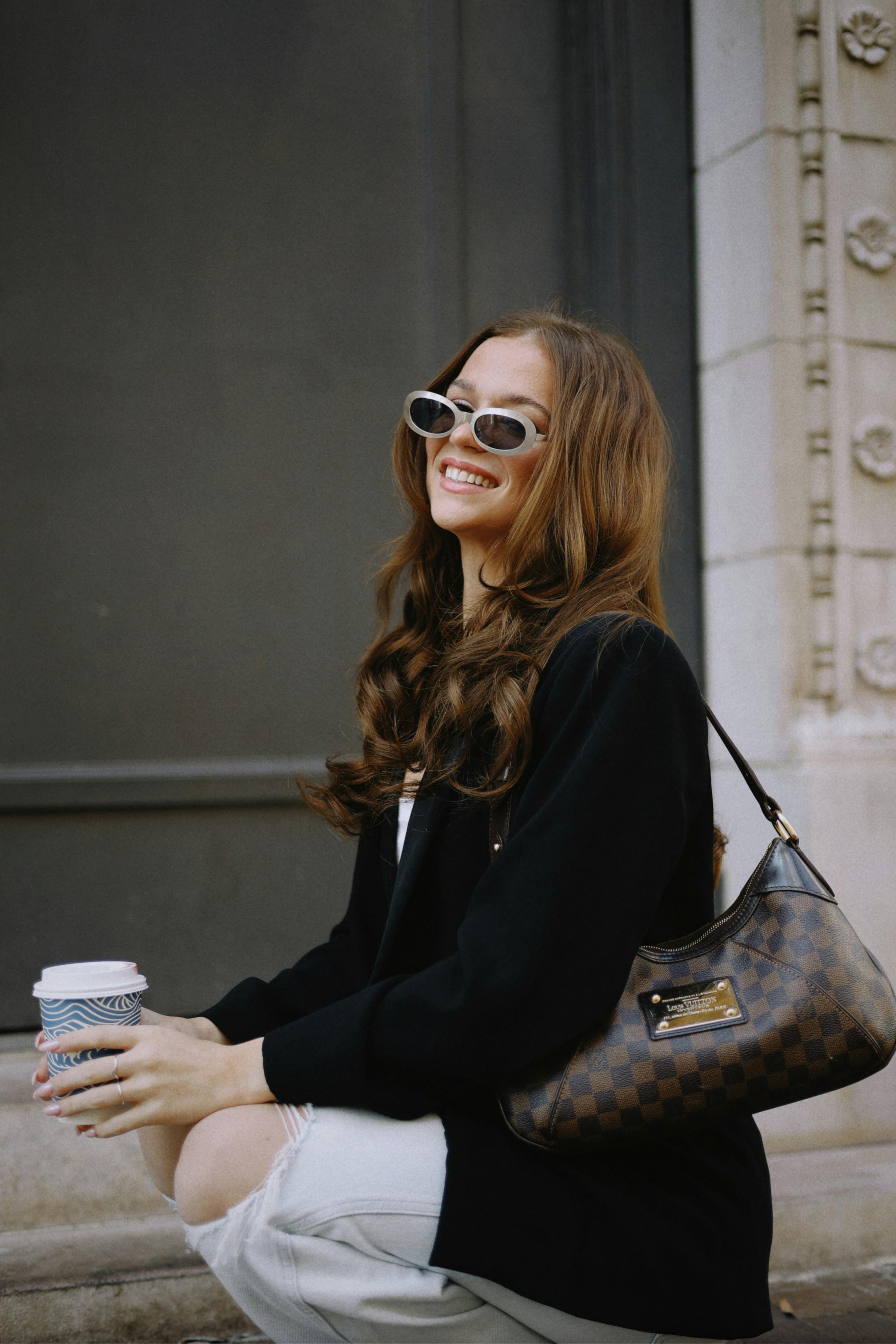 Fashionable woman in sunglasses holding a coffee cup outdoors with a smile.