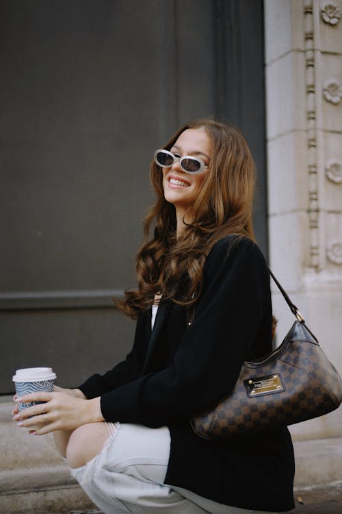Smiling Woman in Sunglasses Crouching with a Coffee Cup in her Hand