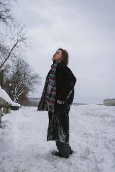 Young woman in winter attire stands amidst snowy landscape, eyes closed, serene expression.