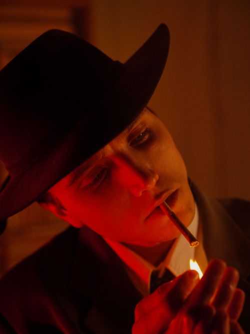 Free A dramatic portrait of a man in a hat lighting a cigarette with red lighting, capturing an elegant ambiance. Stock Photo