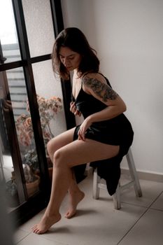 A woman in black satin lingerie sitting on a white stool by a window, with visible tattoos.