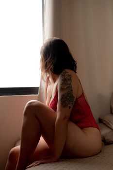 A woman in red lingerie sits on a bed, gazing out a window, showcasing her arm tattoos.