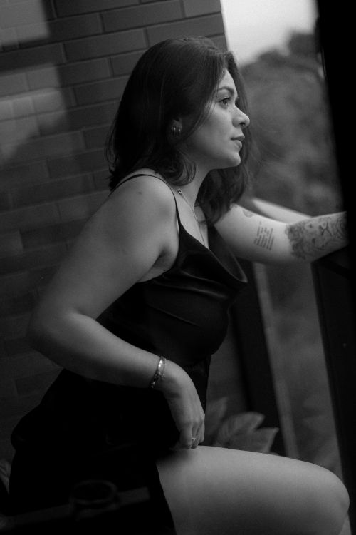 Free A woman in a black dress poses thoughtfully by a window, captured in a black and white portrait. Stock Photo