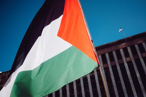 Free A Palestinian flag waving proudly against a blue sky with a modern building backdrop. Stock Photo