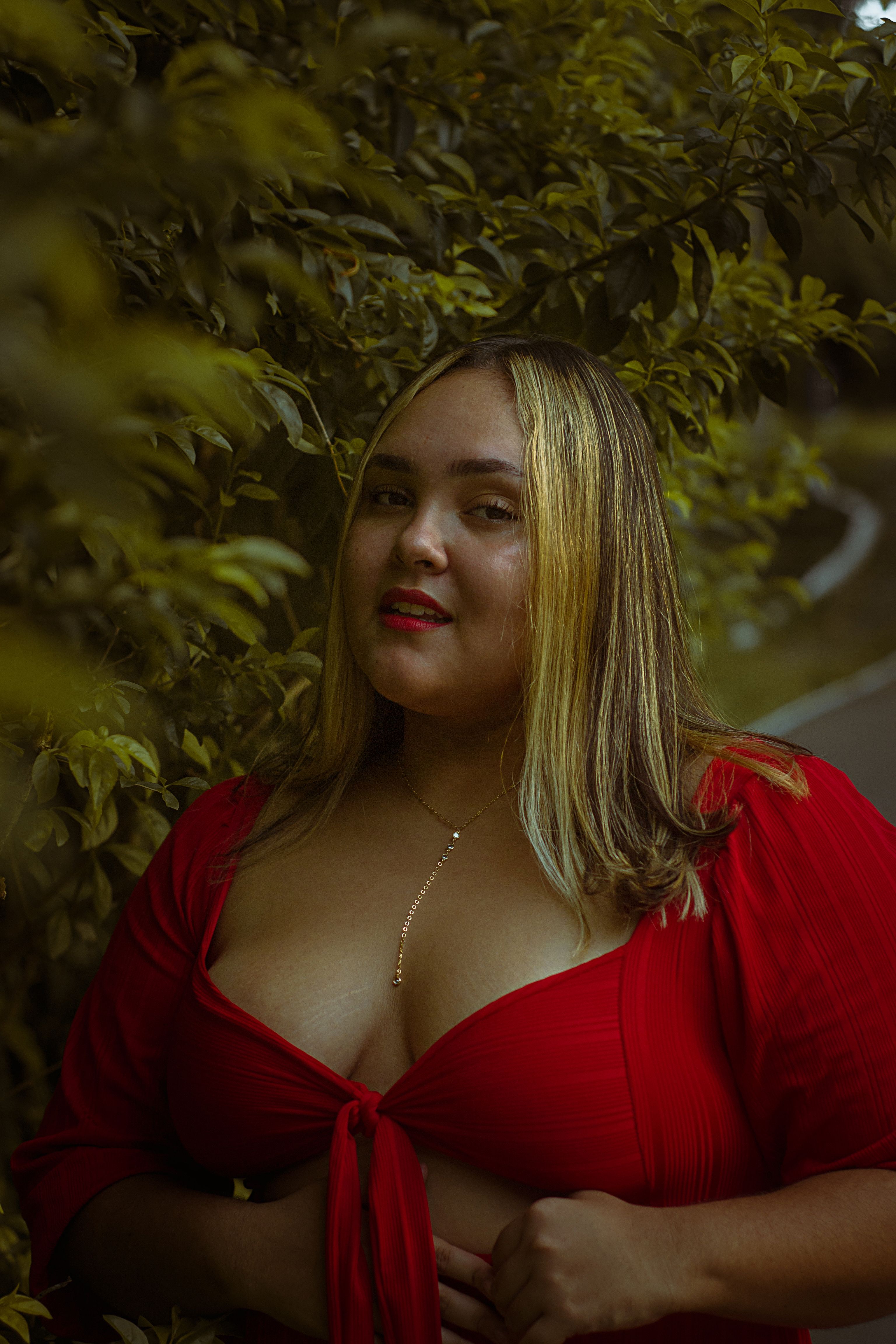 Beautiful woman in a red blouse posing confidently outdoors surrounded by lush greenery.