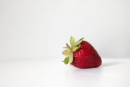 Close-up of a ripe strawberry with green leaves on a minimalist white surface.