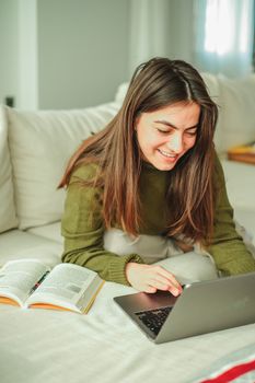 A young woman sits on a sofa working on a laptop, surrounded by books, creating a cozy study environment.