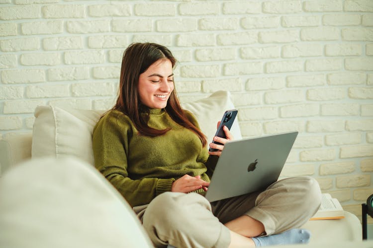 Smiling Woman With A MacBook On Her Lap Texting On A Smartphone While Sitting In The Living Room