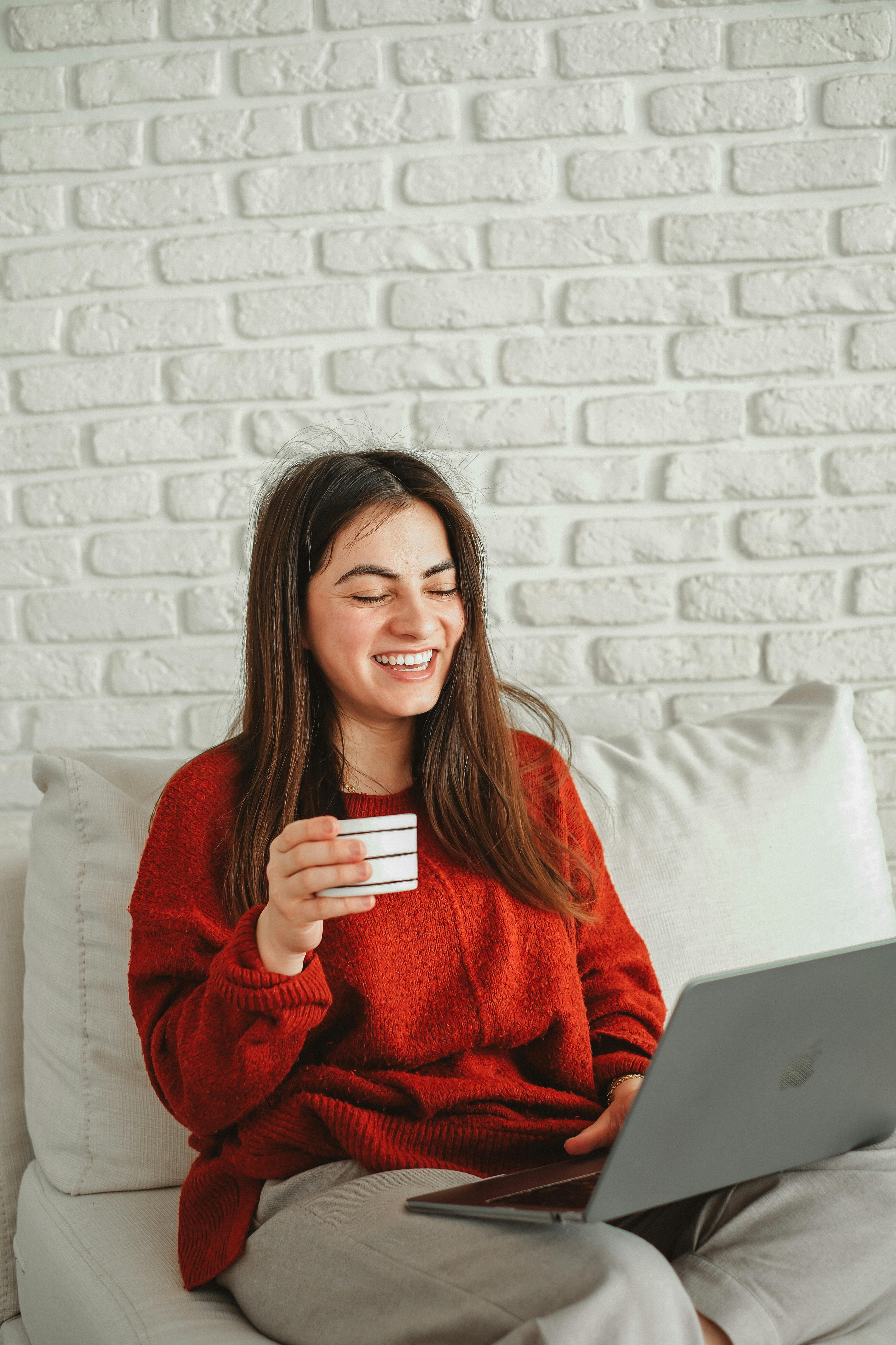 Relaxed woman in red sweater laughs while holding coffee and using laptop indoors.